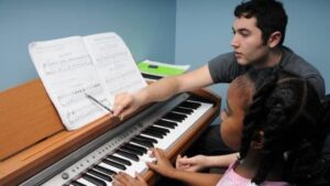 Menina toca piano enquanto é orientada por professor de música. Acima do piano, há uma partitura sendo lida pela menina.