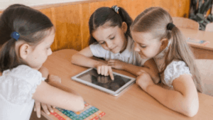 Três meninas brincam com um tablet, sentadas à mesa.