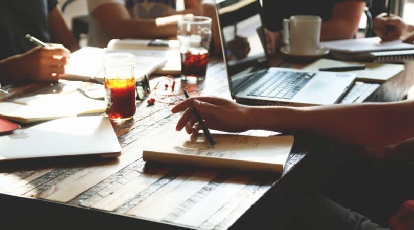 Grupo de estudantes jovens faz trabalho escolar ou de faculdade ao redor de uma mesa com livros, cadernos e computadores. A pesquisa científica representa o objetivo principal da Revista da Escola, Professor, Educação e Tecnologia.